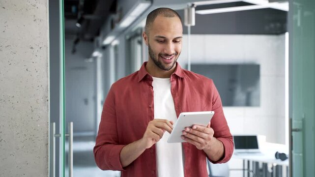 Smiling businessman is using digital tablet while standing in a modern business office. Happy handsome male worker is chatting online, working in app, browsing social media or reading writing message