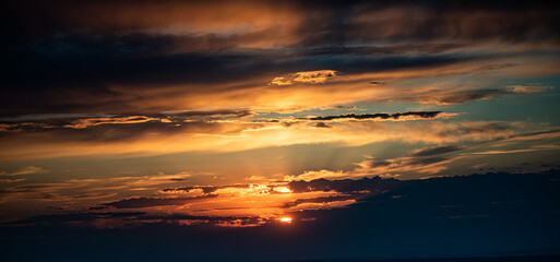 The photo captures a sunset with bright orange and golden sunrays breaking through layered clouds. The horizon is dark, and the sky is filled with dramatic lighting and rich colors.