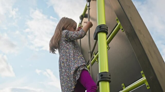 Low-angle view of smiling school age girl climbs a rope ladder on a modern playground. Concept of achievement, fun, freedom and childhood confidence. Outdoor play and active lifestyle.