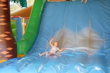 Child joyfully slides down a large inflatable slide in a play area filled with colorful structures and toys
