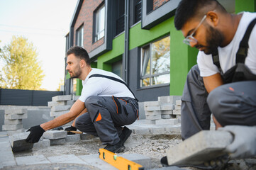 A group of multiracial workers lay paving stones on a sidewalk near a house