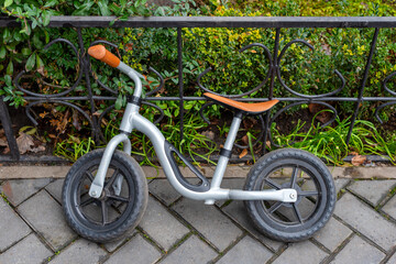 A compact metal balance bike for children parked against a wrought iron fence in a neatly landscaped area with paving and greenery.