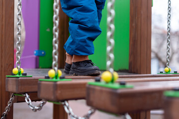 Child walking on playground bridge with chains and colorful wooden elements.