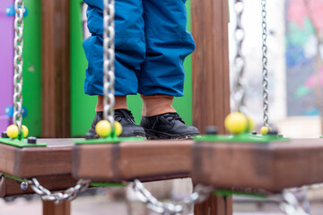 Child walking on playground bridge with chains and colorful wooden elements.
