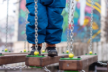 Child walking on playground bridge with chains and colorful wooden elements.