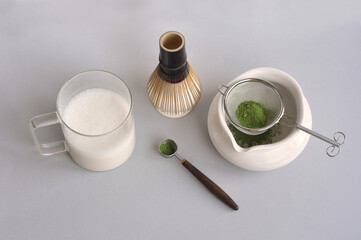 Japanese matcha tea preparation with ceremony traditional tools: white ceramic bowl chawan, glass cup with milk, wooden scoop chashaku, bamboo whisk chasen, matcha powder on grey background.