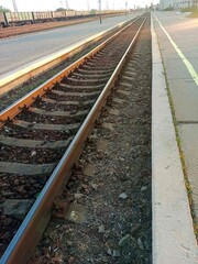 Curved railway tracks at a quiet train station during sunset. The scene features multiple railway lines, overhead electric wires, and a deserted platform with a few distant pedestrians. The warm