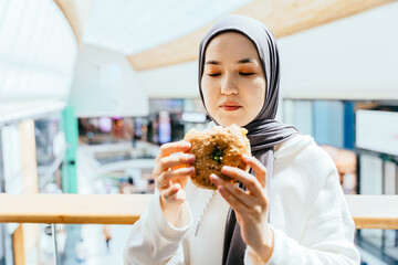 Calm shopper in hijab eating burger near glass railing