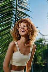 smiling woman with curly hair wearing white crop top outdoors under sunny sky by tropical palm...