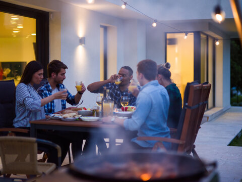 A group of young diverse people having dinner on the terrace of a modern house in the evening. Fun for friends and family. Celebration of holidays, weddings with barbecue - Powered by Adobe