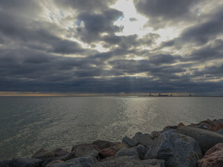 Silty seashore, on the horizon the water meets the dark sky, morning. Sun rays are breaking through the clouds.