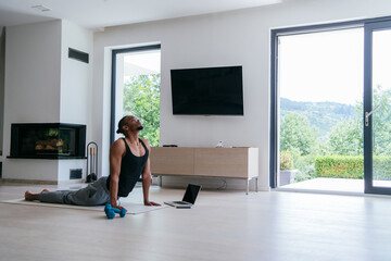Training At Home. Sporty african american man doing the training while watching an online tutorial on a laptop, exercising in the living room, free space. 