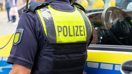 German police officer seen from behind in a yellow vest with "Polizei" text, standing by patrol car. Suitable as background for  breaking news, reports, or public safety and law enforcement topics.