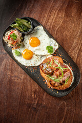Rustic breakfast with sunny-side-up eggs, crispy bacon, hash browns, grilled mushrooms, and herbs on a dark plate over wooden table, top view.
