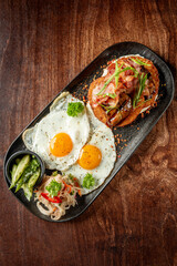 Rustic breakfast with sunny-side-up eggs, crispy bacon, hash browns, grilled mushrooms, and herbs on a dark plate over wooden table, top view.