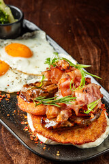 Rustic breakfast with sunny-side-up eggs, crispy bacon, hash browns, grilled mushrooms, and herbs on a dark plate over wooden table, top view.