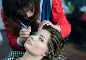 Makeup artist applying eyeliner to a young woman with hair curlers during a beauty session....