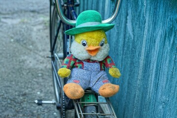 one toy soft plush baby in a green hat funny small with black eyes and an orange beak multi-colored in gray pants beautiful duckling lies on an iron bicycle rack near a wooden wall