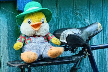 one toy soft plush baby in a green hat funny small with black eyes and an orange beak multi-colored in gray pants beautiful duckling lies on an iron bicycle rack and  saddle near a wooden wall