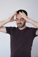 A young man in a casual t-shirt holds his head in both hands with a distressed expression, eyes...