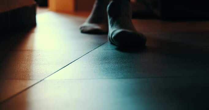 Low-angle close-up of feet in socks walking through a dark room at night. A moody, cinematic shot creating a sense of mystery, suspense, or loneliness.