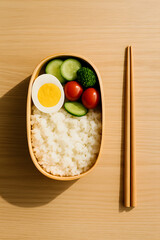 A wooden bento box filled with rice, cucumbers, tomatoes, broccoli, and a boiled egg, served with chopsticks..