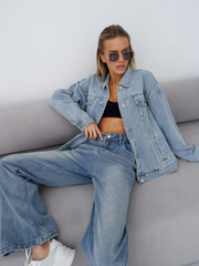 Stylish woman in denim jacket and jeans sitting on sofa, wearing sunglasses and black top. Casual...