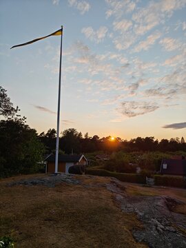 A golden sunset on the Swedish countryside, with a traditional flagpole flying a Swedish pennant beside classic red cottages. Peaceful summer evening with golden light and scenic rural landscape.