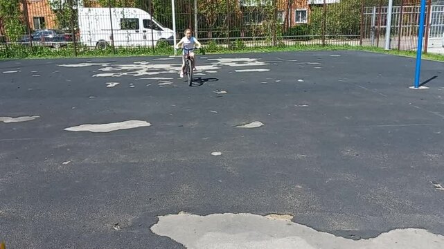 a nine year old girl rides a bicycle on cracked asphalt at a city playground, a boy with a ball runs across the road