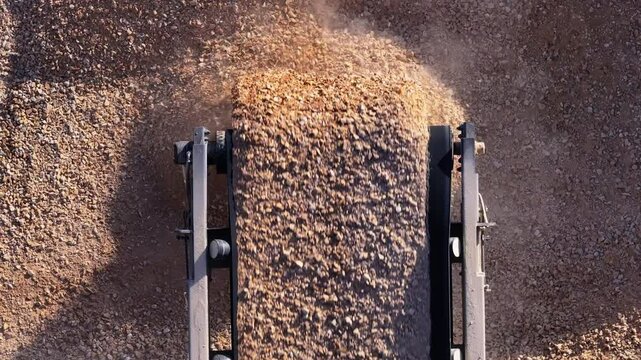 Close-up, top-down aerial of a conveyor belt dropping a continuous stream of crushed stone onto a large stockpile. Seamless loop of industrial aggregate production.
