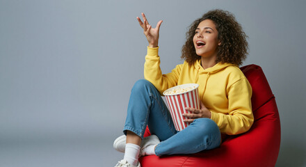 Cheerful young woman enjoying popcorn while sitting on a red bean bag chair, having fun and relaxing at home.