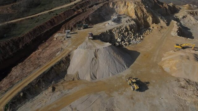 High aerial view of a wheel loader driving to the top of a massive gravel stockpile at a stone quarry, with other industrial machinery working in the background.
