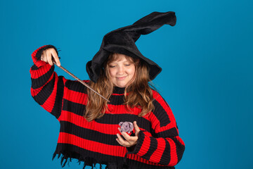 Halloween Theme. Young witch girl in striped sweater and black hat holding magic stick, toy mouse and making spell against blue background.