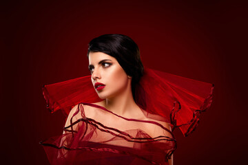 Tempting young woman with red veiled dress posing against red gradient background, portraying...