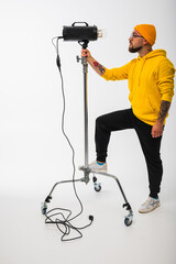 Smiling man in yellow hoodie holding professional lighting equipment in studio. Behind-the-scenes concept with cheerful crew member during photo or video shoot setup