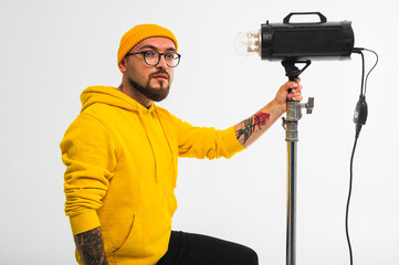 Smiling man in yellow hoodie holding professional lighting equipment in studio. Behind-the-scenes concept with cheerful crew member during photo or video shoot setup