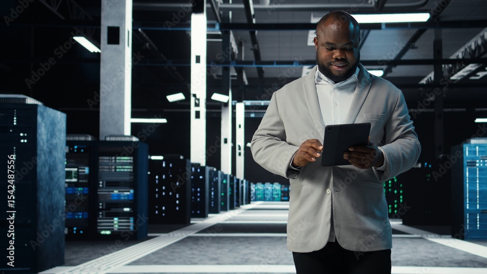 Poster system administrator walking in data center analyzing infrastructure performance outputs on tablet. 