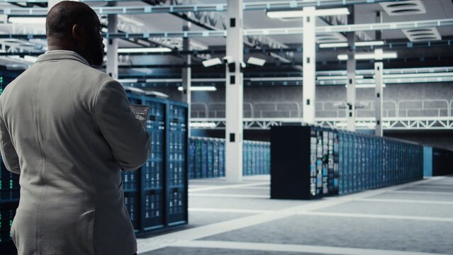 Engineer walking in data center using device to configure hardware optimized for AI tasks. Man doing maintenance on rigs performing load balancing for artificial intelligence computing, camera A