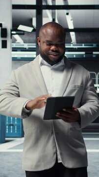 Vertical video Smiling admin using tablet in in data center maintaining infrastructure for workloads. Joyful IT professional in server farm utilizing high tech server clusters for parallel processing