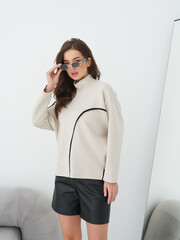 Woman adjusts sunglasses, wearing beige turtleneck with black accents and leather shorts, while...