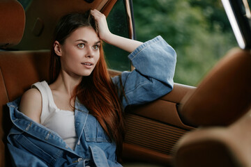 Young woman with long hair in denim jacket, sitting in a car, looking thoughtful and relaxed, with...