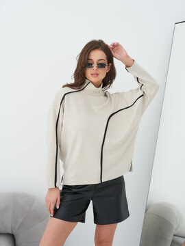 Woman with wavy brown hair wears beige sweater with black lines, black shorts, and sunglasses, posing confidently. Mirror reflects part of her arm. Small couch visible in corner