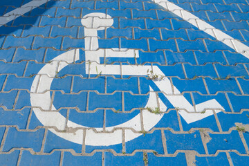 White wheelchair parking symbol on blue interlocking pavement, with grass growing between tiles. Urban accessibility detail