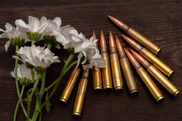 A bouquet of white chrysanthemums lies next to rifle cartridges on a dark wooden surface. The image symbolises the contrast between peace and war, life and death, or the memory of the fallen.