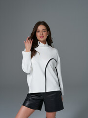 Female model poses in white turtleneck sweater with black trim, paired with black leather shorts. Her long hair is loose, and one hand is raised near her face, expressing elegance