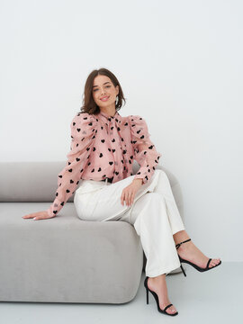Smiling woman sits on modern gray sofa wearing pink blouse with black heart pattern and white wide-leg pants. Her black high heels add elegance. Long brown hair frames her face