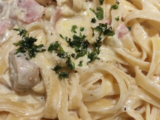 A close-up image of creamy pasta, likely fettuccine Alfredo, topped with chopped parsley. The dish includes mushrooms, bits of ham or bacon, and is coated in a rich white sauce with grated cheese.