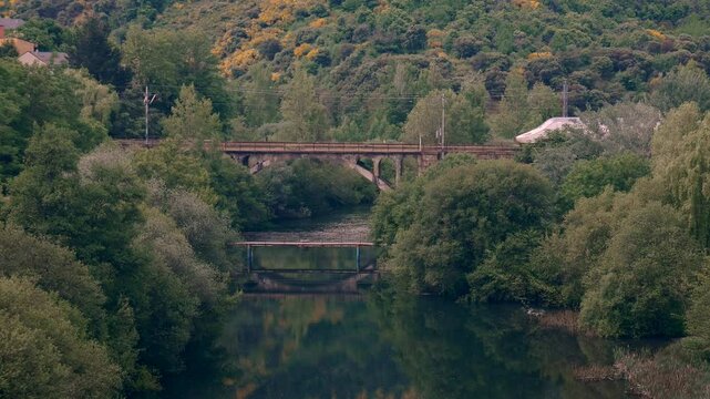 The Renfe Alvia train of the Spanish public operator Renfe Viajeros leaves Ponferrada station, passing over a bridge and leaving its reflection in the Sil River