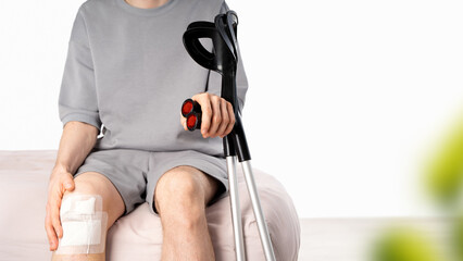 Male person with injury sits holding elbow crutches beside bandaged leg, bruising and limited mobility. Concept of orthopedic trauma, domestic healing, physical recovery process. Banner. Copy space