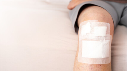 Detailed shot of male leg with visible adhesive bandage after knee trauma on soft neutral bedding. Concept of home wound recovery, injury treatment and protective care. Banner. Copy space